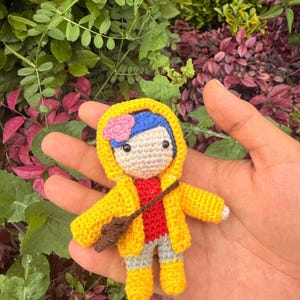 May include: A small crochet doll featuring blue hair with a pink flower, wearing a yellow hooded raincoat, a red shirt, gray pants, and yellow boots. The doll has a brown crossbody bag and black bead eyes, crafted with detailed yarn stitching.
