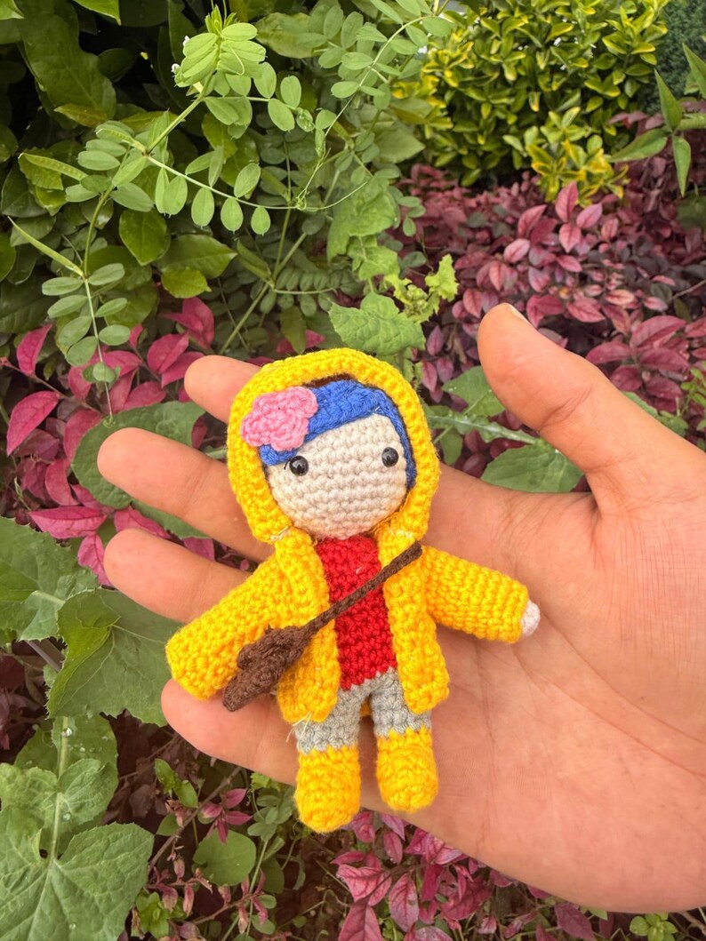 May include: A small crochet doll featuring blue hair with a pink flower, wearing a yellow hooded raincoat, a red shirt, gray pants, and yellow boots. The doll has a brown crossbody bag and black bead eyes, crafted with detailed yarn stitching.