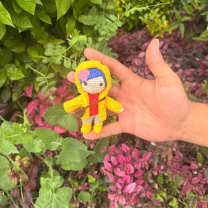 May include: A small crocheted doll featuring blue hair with a pink flower accessory, wearing a bright yellow hooded raincoat over a red shirt and light-colored pants, and carrying a small brown cross-body bag across its chest.