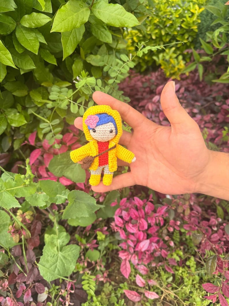May include: A small crocheted doll featuring blue hair with a pink flower accessory, wearing a bright yellow hooded raincoat over a red shirt and light-colored pants, and carrying a small brown cross-body bag across its chest.