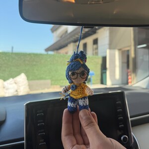 Pode incluir: Uma pequena boneca de croch&ecirc; feita &agrave; m&atilde;o com cabelo azul, &oacute;culos e uma parte superior amarela. A boneca est&aacute; usando cal&ccedil;as listradas azuis e brancas. Est&aacute; pendurada por um fio, provavelmente como acess&oacute;rio de carro.