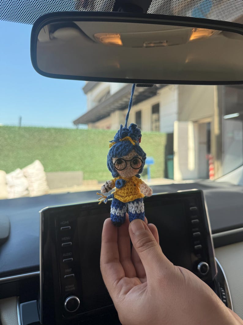 Pode incluir: Uma pequena boneca de croch&ecirc; feita &agrave; m&atilde;o com cabelo azul, &oacute;culos e uma parte superior amarela. A boneca est&aacute; usando cal&ccedil;as listradas azuis e brancas. Est&aacute; pendurada por um fio, provavelmente como acess&oacute;rio de carro.