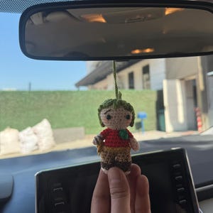 Pode incluir: Boneca de croch&ecirc; feita &agrave; m&atilde;o com chap&eacute;u verde, camisa vermelha e cal&ccedil;as marrons. A boneca tem um pequeno detalhe de folha verde na camisa e &eacute; suspensa por uma corda verde. A boneca &eacute; segurada em frente ao painel e ao espelho retrovisor de um carro.