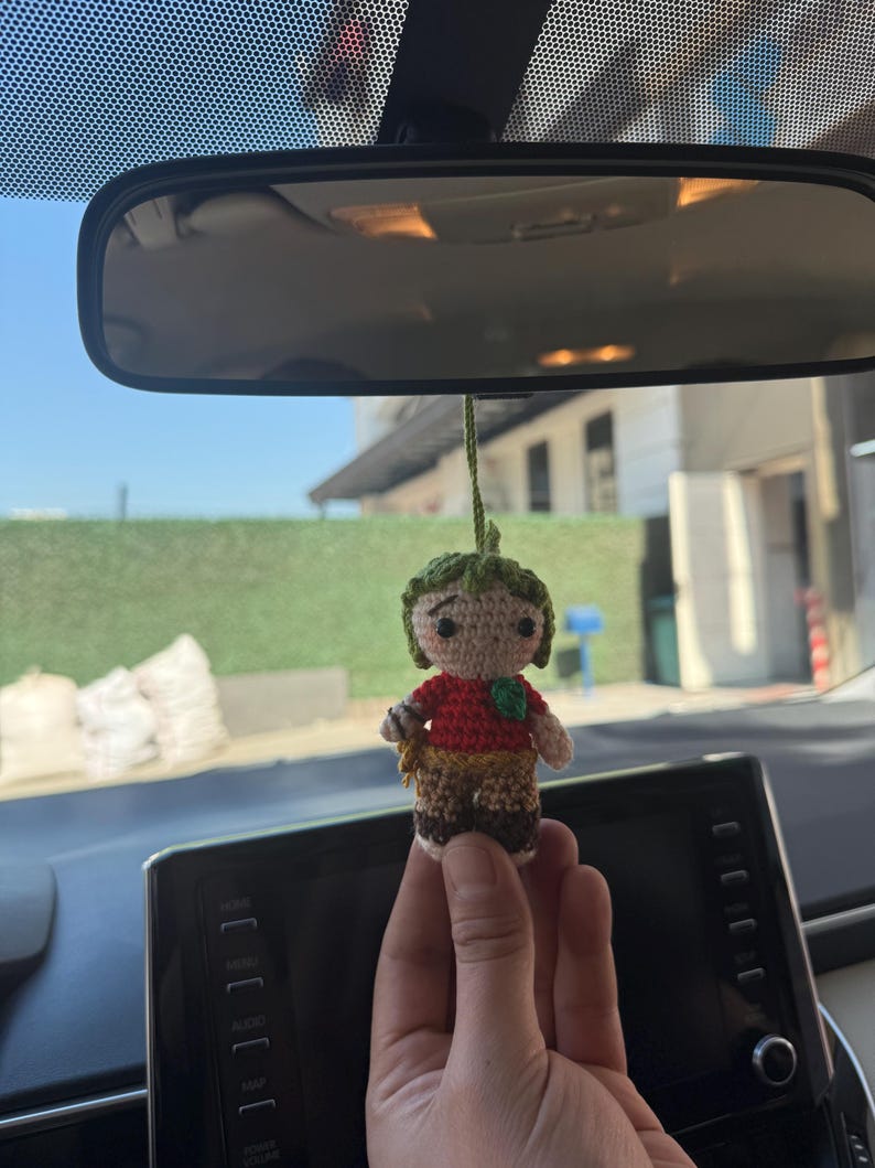 Pode incluir: Boneca de croch&ecirc; feita &agrave; m&atilde;o com chap&eacute;u verde, camisa vermelha e cal&ccedil;as marrons. A boneca tem um pequeno detalhe de folha verde na camisa e &eacute; suspensa por uma corda verde. A boneca &eacute; segurada em frente ao painel e ao espelho retrovisor de um carro.