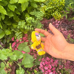 May include: A small crocheted doll featuring a bright yellow hooded outfit, a brown satchel across the chest, and light-colored yarn details on the face and feet. The doll is crafted with visible yarn stitches and has a compact, stuffed construction.