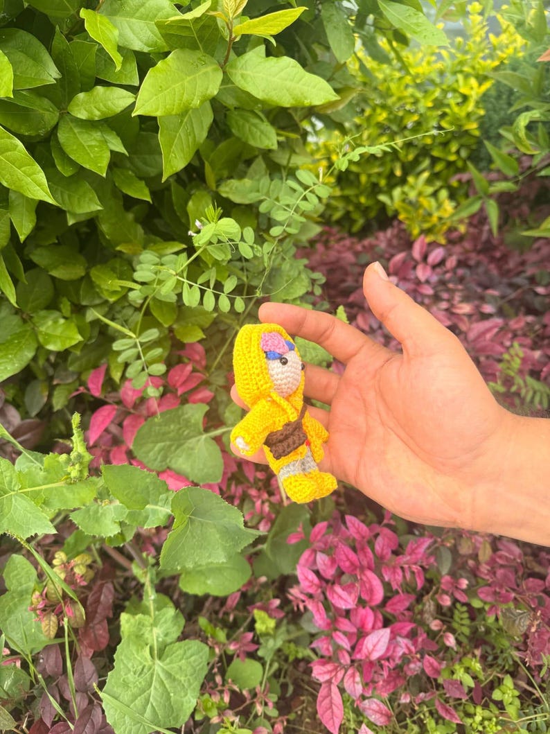 May include: A small crocheted doll featuring a bright yellow hooded outfit, a brown satchel across the chest, and light-colored yarn details on the face and feet. The doll is crafted with visible yarn stitches and has a compact, stuffed construction.