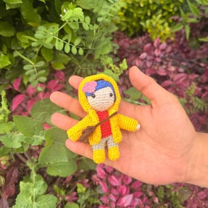 May include: A small crocheted doll featuring blue hair with a pink flower accessory, wearing a bright yellow hooded raincoat, a red shirt, gray pants, and yellow boots. The doll has a brown crossbody bag and a light beige face with black eyes.