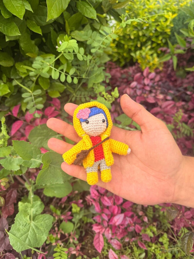 May include: A small crocheted doll featuring blue hair with a pink flower accessory, wearing a bright yellow hooded raincoat, a red shirt, gray pants, and yellow boots. The doll has a brown crossbody bag and a light beige face with black eyes.