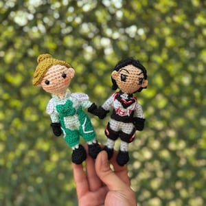 Op de afbeelding: Twee gehaakte poppen, een in groen en wit, de andere in grijs en rood, houden elkaars hand vast. De poppen hebben zwarte schoenen en staan tegen een vage groene achtergrond. De poppen zijn handgemaakt en hebben gedetailleerde gezichtskenmerken.