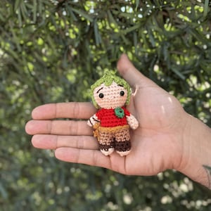 Pode incluir: Boneca de croch&ecirc; feita &agrave; m&atilde;o com cabe&ccedil;a verde folhosa, camisa vermelha e cal&ccedil;as marrons. A boneca tem uma express&atilde;o neutra e &eacute; segurada em uma m&atilde;o aberta contra um fundo verde desfocado. A boneca tem aproximadamente 7,6 cm de altura.