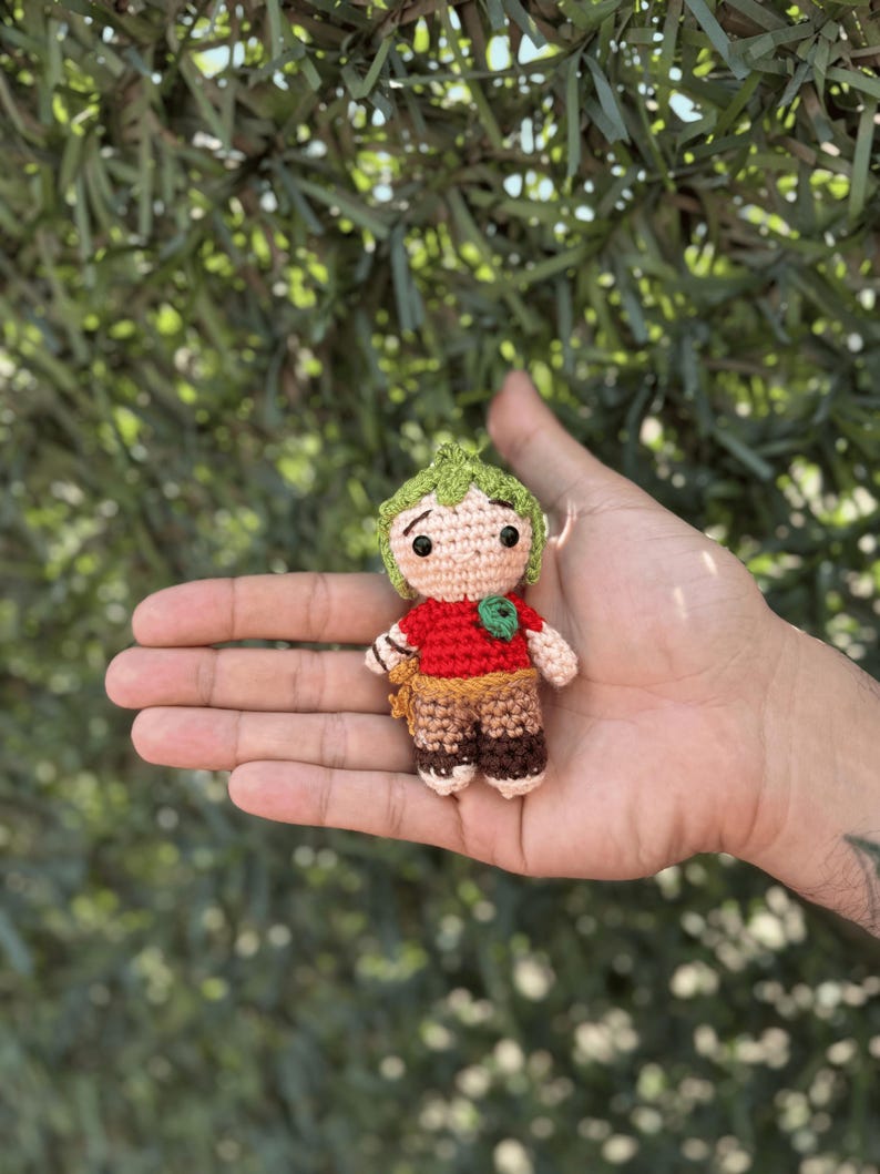 Pode incluir: Boneca de croch&ecirc; feita &agrave; m&atilde;o com cabe&ccedil;a verde folhosa, camisa vermelha e cal&ccedil;as marrons. A boneca tem uma express&atilde;o neutra e &eacute; segurada em uma m&atilde;o aberta contra um fundo verde desfocado. A boneca tem aproximadamente 7,6 cm de altura.