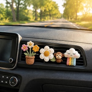 May include: A set of three crochet car air vent clips featuring a potted plant with pink, white, and yellow flowers, a large white daisy with a yellow center next to a small brown monkey, and a white cloud with a colorful rainbow hanging below.
