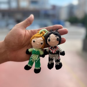 May include: Two small, handmade crochet dolls. One doll has blonde hair and wears a green outfit, the other has black hair and a black and gray outfit. Both dolls have black shoes and are attached to a keychain.