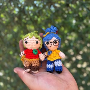 以下が含まれることがあります： 2つの手作りのかぎ針編み人形。1つは赤いシャツ、茶色のズボン、緑の髪をしています。もう1つは黄色のシャツ、青いズボン、メガネ、青い髪をしています。両方の人形は手をつないでいます。