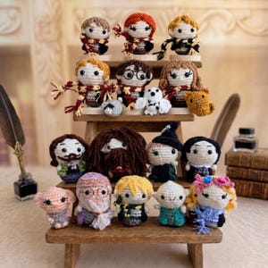 May include: A collection of small, handcrafted crochet dolls representing various characters. The figures feature yarn hair, embroidered facial details, and miniature accessories like scarves, glasses, and hats, arranged on a tiered wooden display stand.