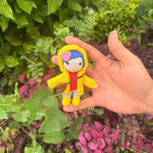May include: A small crocheted doll featuring blue hair with a pink flower, wearing a bright yellow hooded raincoat, a red shirt, and light-colored pants. The doll has a brown crossbody bag and yellow boots, crafted with detailed yarn stitching.