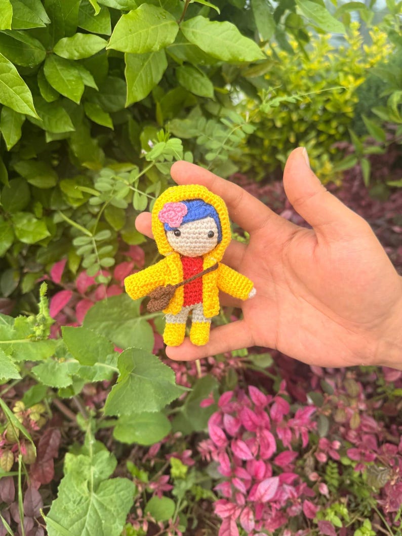 May include: A small crocheted doll featuring blue hair with a pink flower, wearing a bright yellow hooded raincoat, a red shirt, and light-colored pants. The doll has a brown crossbody bag and yellow boots, crafted with detailed yarn stitching.