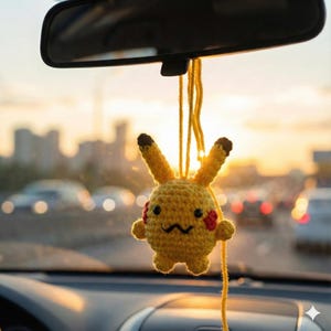 Może przedstawiać: Żółty szydełkowy urok do samochodu Pikachu, zawieszony na lusterku wstecznym samochodu. Urok ma czarne oczy, zygzakowate usta, czerwone policzki i uszy z brązowymi końcówkami. Tło przedstawia panoramę miasta i zachód słońca.