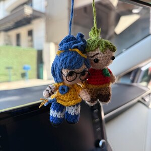 Pode incluir: Duas bonecas de croch&ecirc; penduradas no espelho retrovisor de um carro. Uma boneca tem cabelo azul, &oacute;culos e uma roupa amarela com listras azuis. A outra boneca tem cabelo verde, uma camisa vermelha e cal&ccedil;as marrons. Ambas as bonecas s&atilde;o feitas &agrave; m&atilde;o e t&ecirc;m uma apar&ecirc;ncia fantasiosa.