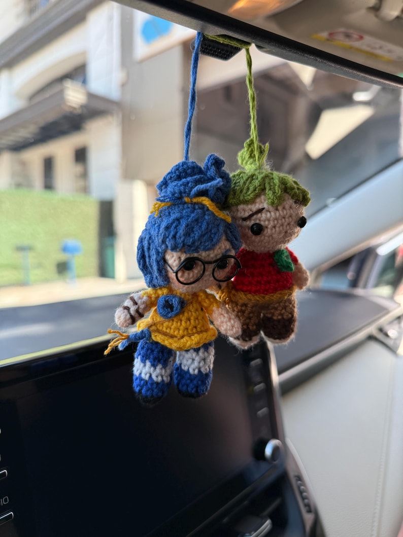 Pode incluir: Duas bonecas de croch&ecirc; penduradas no espelho retrovisor de um carro. Uma boneca tem cabelo azul, &oacute;culos e uma roupa amarela com listras azuis. A outra boneca tem cabelo verde, uma camisa vermelha e cal&ccedil;as marrons. Ambas as bonecas s&atilde;o feitas &agrave; m&atilde;o e t&ecirc;m uma apar&ecirc;ncia fantasiosa.
