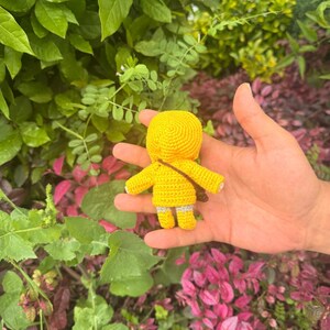 May include: A small, handmade crochet doll featuring a bright yellow hooded raincoat, yellow boots with grey cuffs, and a thin brown strap worn diagonally across the back. The doll is crafted with a dense, textured yarn stitch pattern throughout.