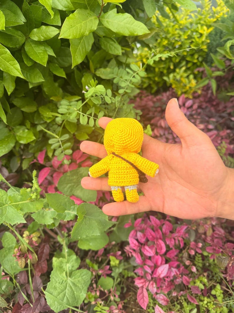 May include: A small, handmade crochet doll featuring a bright yellow hooded raincoat, yellow boots with grey cuffs, and a thin brown strap worn diagonally across the back. The doll is crafted with a dense, textured yarn stitch pattern throughout.