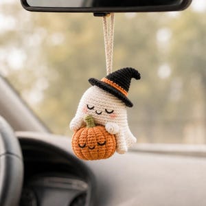 May include: A crocheted hanging ornament featuring a small white ghost with pink cheeks wearing a black witch hat with an orange band, holding a round orange pumpkin with a green stem. Both the ghost and pumpkin have embroidered smiling faces.