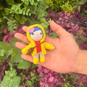 May include: A small crocheted doll featuring a yellow hooded raincoat, a red shirt, gray pants, and yellow boots. The doll has blue hair adorned with a small pink flower and carries a brown crossbody bag. The figure is crafted with yarn.