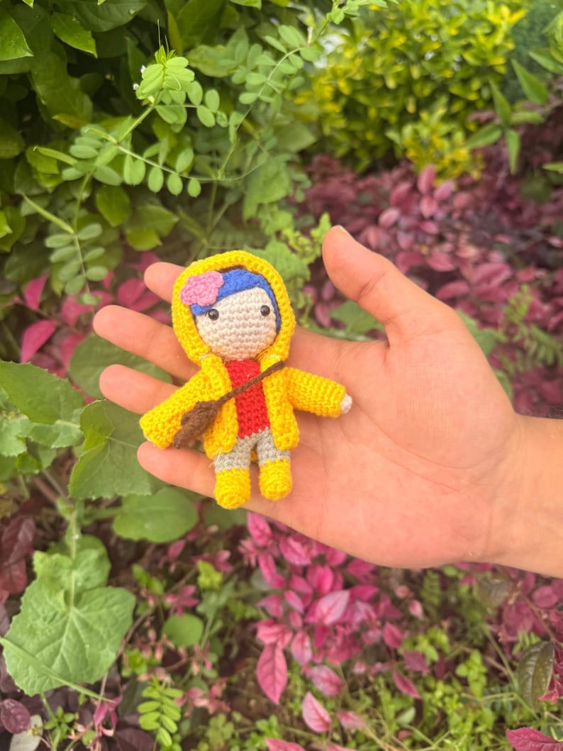 May include: A small crocheted doll featuring a yellow hooded raincoat, a red shirt, gray pants, and yellow boots. The doll has blue hair adorned with a small pink flower and carries a brown crossbody bag. The figure is crafted with yarn.