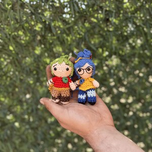 Pode incluir: Duas pequenas bonecas de croch&ecirc; feitas &agrave; m&atilde;o. Uma boneca tem cabelo verde, uma camisa vermelha e cal&ccedil;as marrons. A outra boneca tem cabelo azul, uma camisa amarela e cal&ccedil;as azuis. Ambas as bonecas est&atilde;o de m&atilde;os dadas. O fundo &eacute; folhagem verde desfocada.