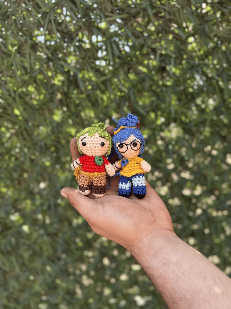 Pode incluir: Duas pequenas bonecas de croch&ecirc; feitas &agrave; m&atilde;o. Uma boneca tem cabelo verde, uma camisa vermelha e cal&ccedil;as marrons. A outra boneca tem cabelo azul, uma camisa amarela e cal&ccedil;as azuis. Ambas as bonecas est&atilde;o de m&atilde;os dadas. O fundo &eacute; folhagem verde desfocada.