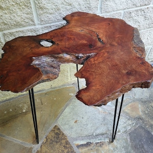 May include: A rustic wooden table with a live edge top and a natural wood finish. The table is supported by three black metal hairpin legs.