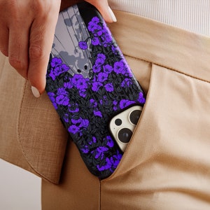 May include: A black phone case with a floral design featuring purple flowers on a black background. The case is in a person's pocket.