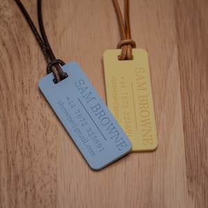 Peut inclure: Deux &eacute;tiquettes rectangulaires de nom, l'une bleue et l'autre jaune, avec un cordon en cuir brun. Le texte sur les &eacute;tiquettes indique "SAM BROWNE" et comprend un num&eacute;ro de t&eacute;l&eacute;phone et une adresse e-mail.