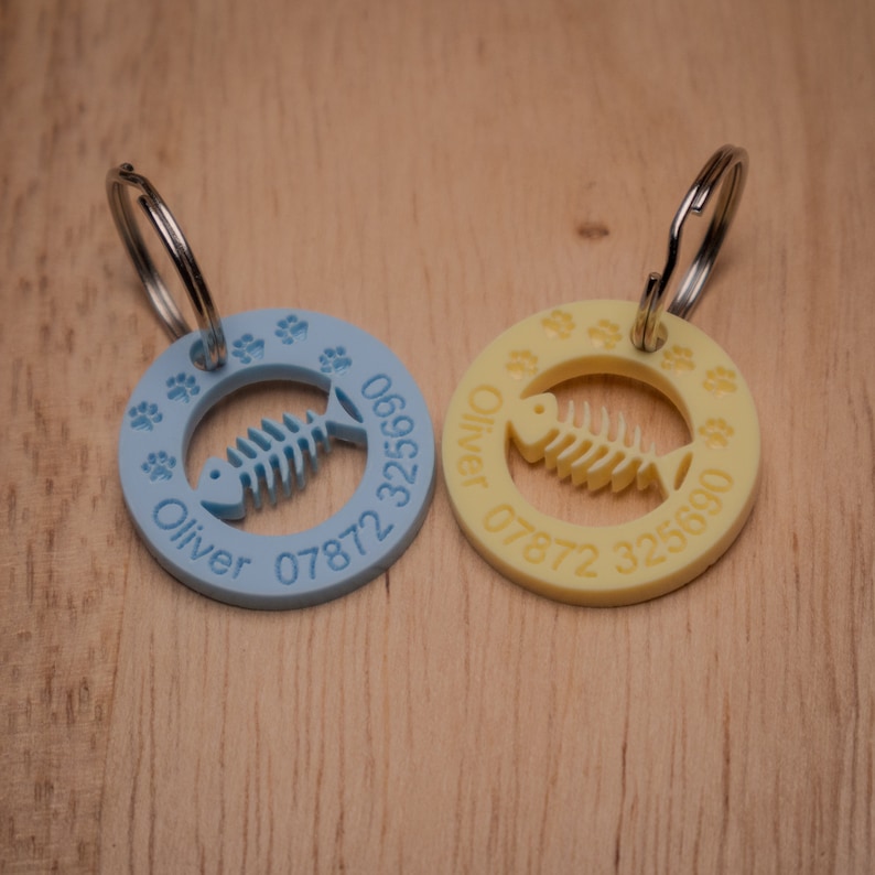 May include: Two personalized pet ID tags, one blue and one yellow, with a fishbone design and paw prints. The tags are engraved with the name "Oliver" and phone numbers.
