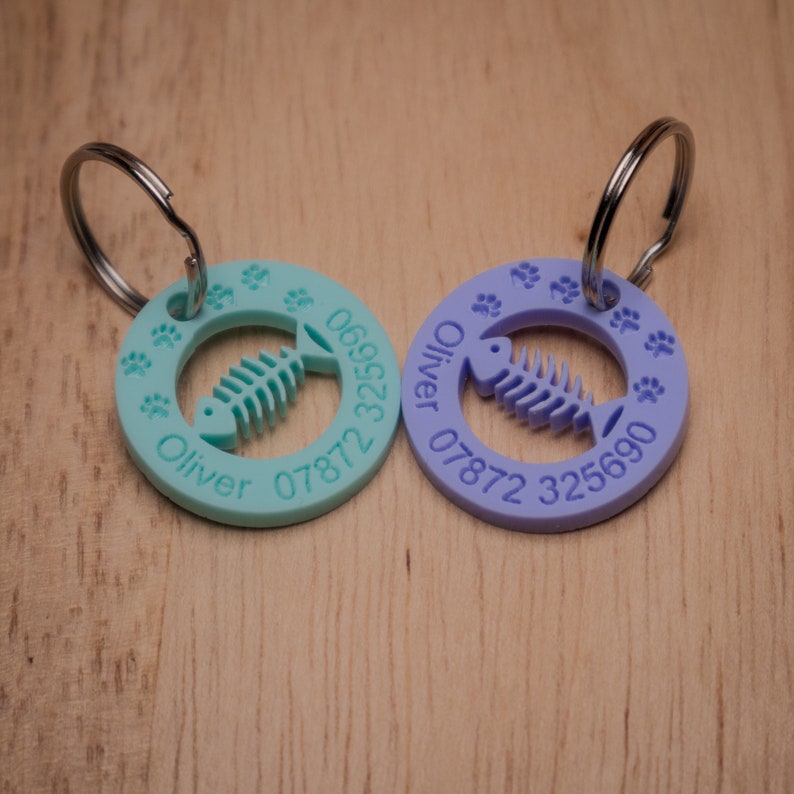 May include: Two round pet ID tags with a fishbone design and paw prints. The tag on the left is turquoise blue with the name "Oliver" and the phone number "06928 07872". The tag on the right is purple with the name "Oliver" and the phone number "07872 325690".