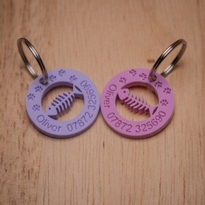 May include: Two purple and pink personalized pet ID tags with a fish bone design and paw prints. The tags are attached to silver key rings. The text on the tags reads "Oliver 07872 069522" and "Oliver 07872 325690".