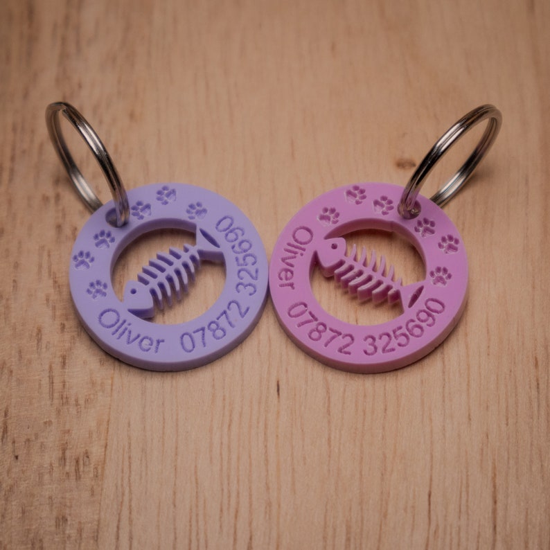 May include: Two purple and pink personalized pet ID tags with a fish bone design and paw prints. The tags are attached to silver key rings. The text on the tags reads "Oliver 07872 069522" and "Oliver 07872 325690".