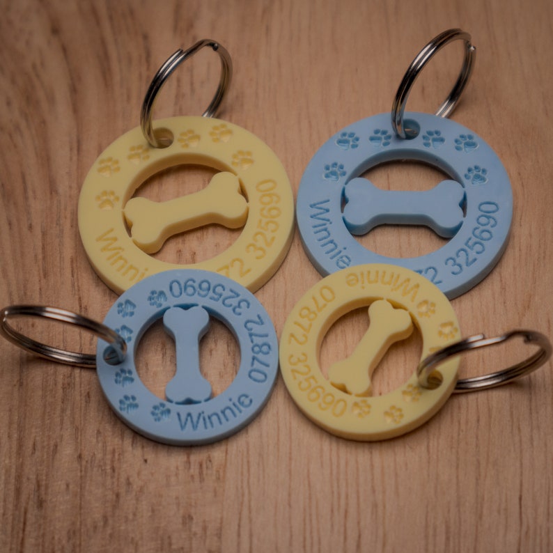 May include: Four yellow and blue dog tags with paw prints and a bone design. The tags have the name "Winnie" and a phone number on them. The tags are attached to silver key rings.
