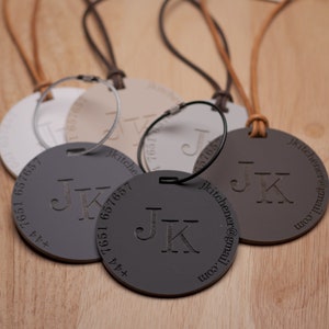 May include: Four round leather keychains with a metal ring. Each keychain has the initials "JK" engraved in the centre and a phone number and email address around the edge. The keychains are in different colours: white, beige, grey, and brown.