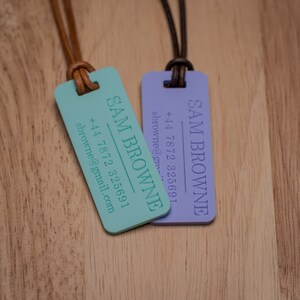 Peut inclure: Deux &eacute;tiquettes rectangulaires turquoise et violettes avec des lani&egrave;res en cuir marron. Les &eacute;tiquettes portent le nom "SAM BROWNE" ainsi qu'un num&eacute;ro de t&eacute;l&eacute;phone et une adresse e-mail.