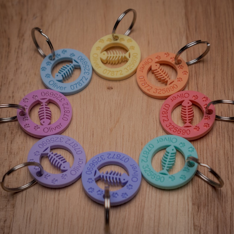 May include: A set of seven colorful cat ID tags with a fishbone design and the name "Oliver" and a phone number. The tags are made of plastic and have a metal ring for attaching to a collar.