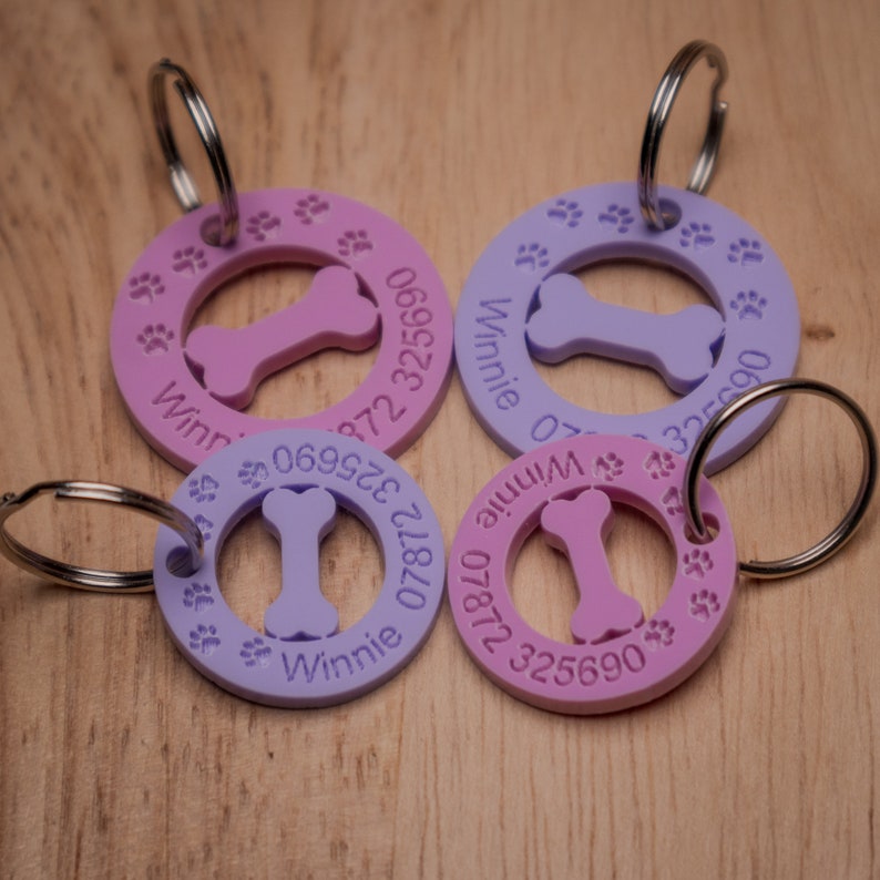 May include: Four round, pink and purple dog tags with a bone shape cut out of the center. Each tag has a name, "Winnie", and a phone number printed on it. The tags are attached to silver key rings.
