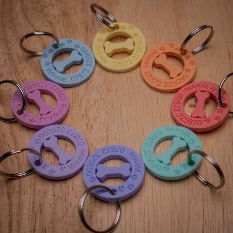 May include: A set of eight colorful dog tag keychains with a bone-shaped cutout in the center. Each tag features a different color and has the name "Winnie" and a phone number printed on it.