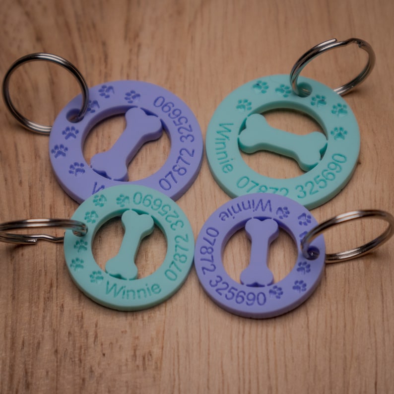 May include: Four round, purple and turquoise dog tags with a bone-shaped cutout and paw prints around the edge. The tags are engraved with the name "Winnie" and phone numbers.