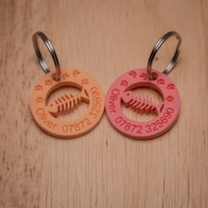 May include: Two orange and pink personalized pet ID tags with a fish bone design and the name Oliver and a phone number.