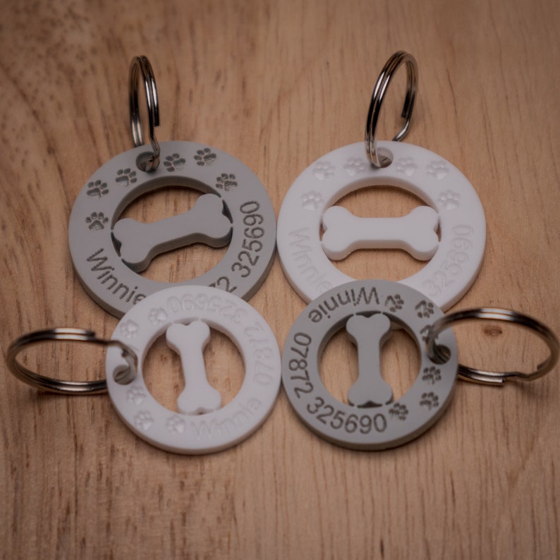 Puede incluir: Cuatro chapas redondas para perros, grises y blancas, con un recorte en forma de hueso y huellas de patas alrededor del borde. Las chapas tienen el nombre "Winnie" y n&uacute;meros de tel&eacute;fono grabados.