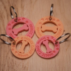 May include: Four pink and orange dog tags with a bone shape cut out in the center. Each tag has the name "Winnie" and a phone number printed on it.