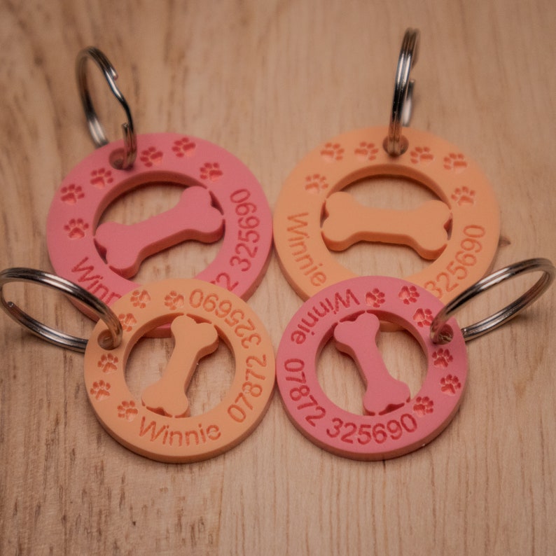 May include: Four pink and orange dog tags with a bone shape cut out in the center. Each tag has the name "Winnie" and a phone number printed on it.