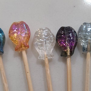 May include: Five colorful, iridescent, glass-like ornaments on wooden sticks. The ornaments are in shades of blue, orange, clear, purple, and silver. Each has a unique, textured design. The sticks are thin and light-colored.